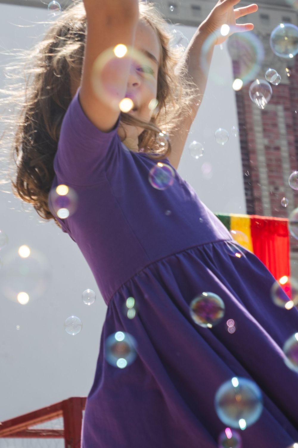 A young girl in a purple dress reaches up to catch colorful soap bubbles floating around her outdoors.