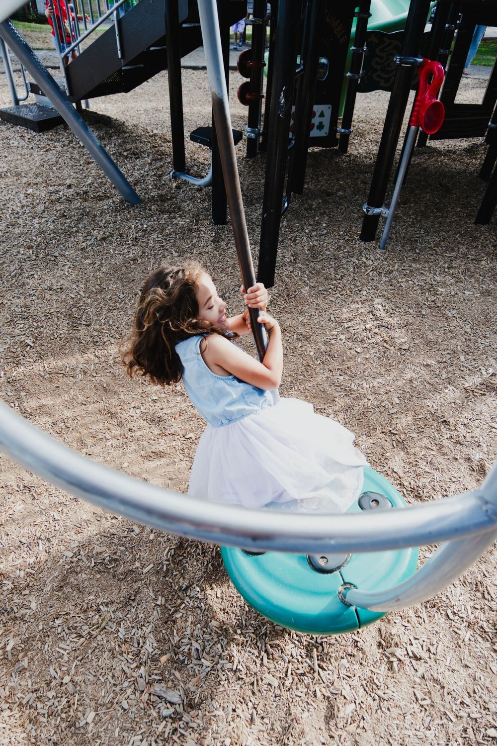 Young girl in white dress riding a disc swing at a playground, smiling and holding the chains.