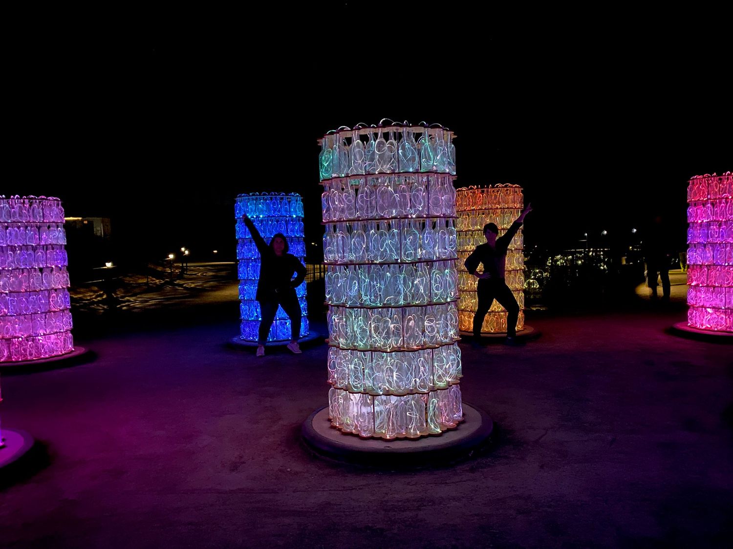 Silhouettes of people stand among glowing cylindrical sculptures made of plastic bottles illuminated in pink and blue at night.