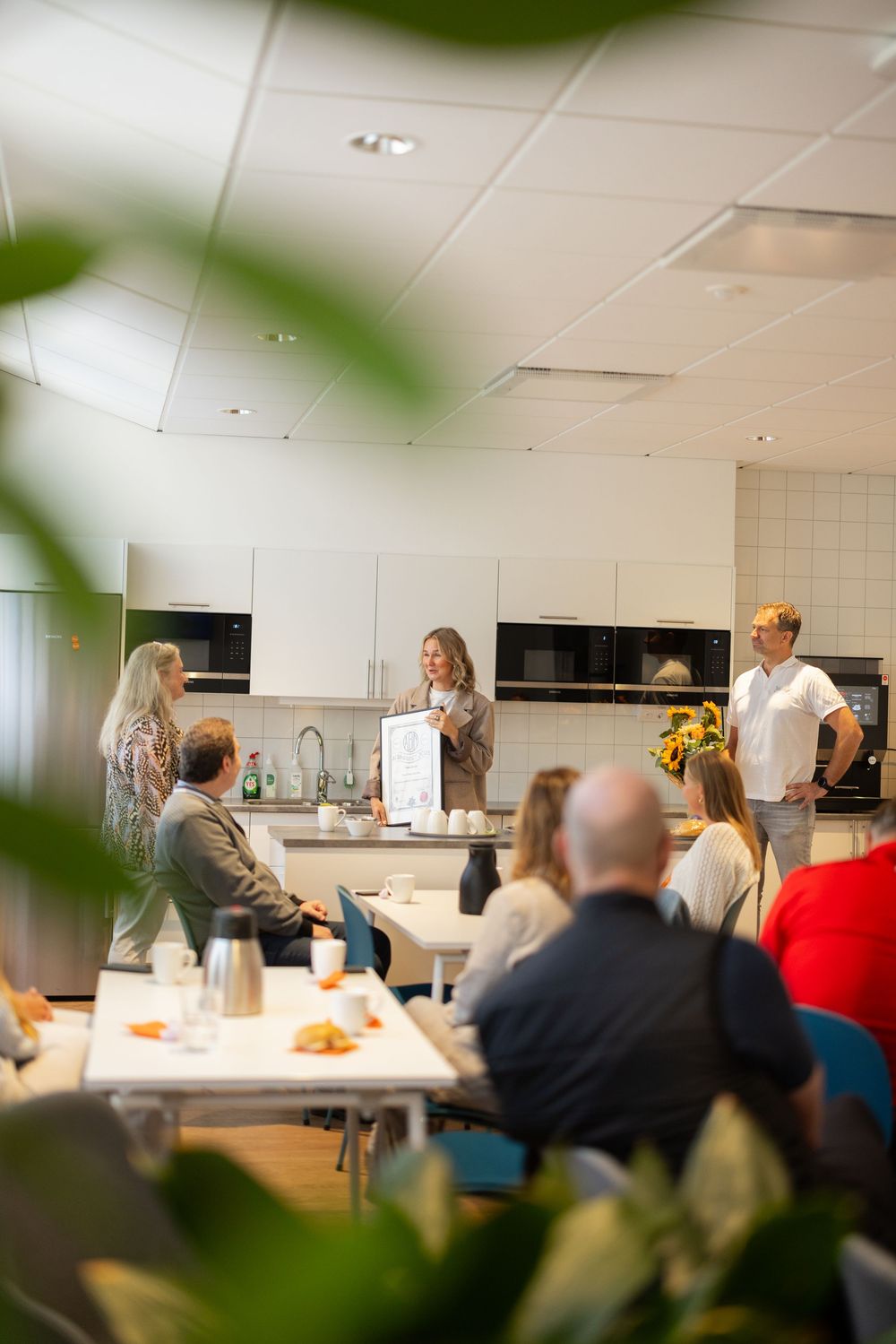 Group of colleagues gathered in a modern office kitchen for a presentation, with a woman speaking at a whiteboard.