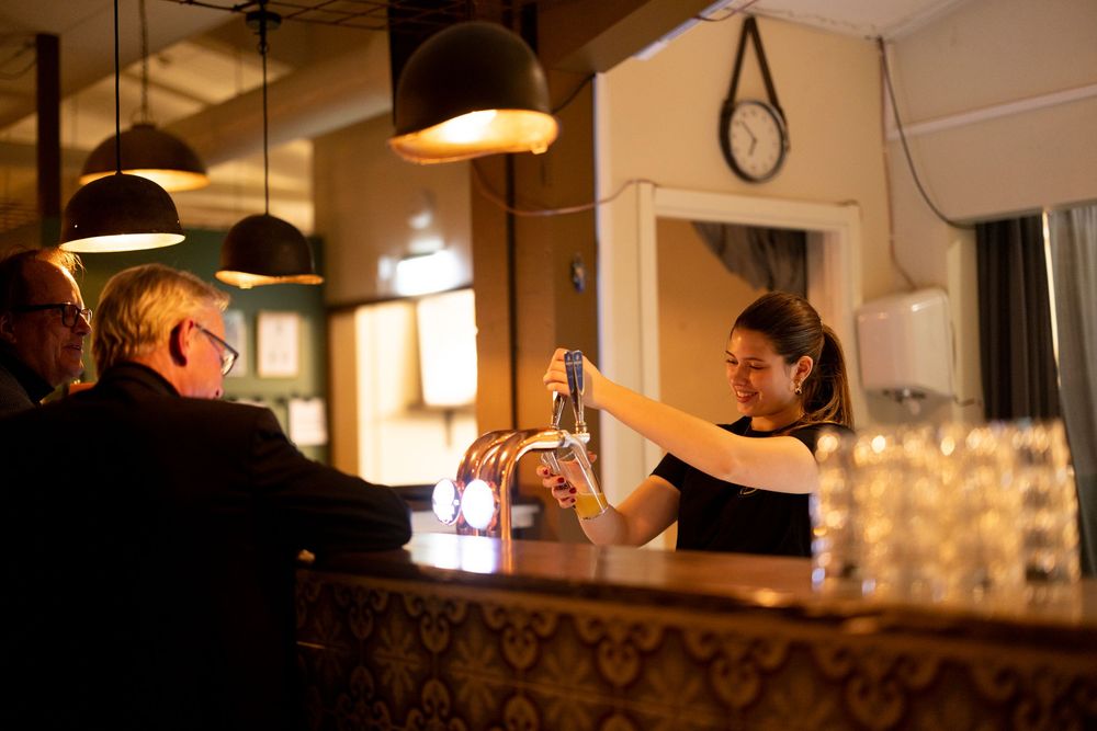 Bartender pouring drinks at a warmly lit bar while serving customers in a cozy restaurant setting.