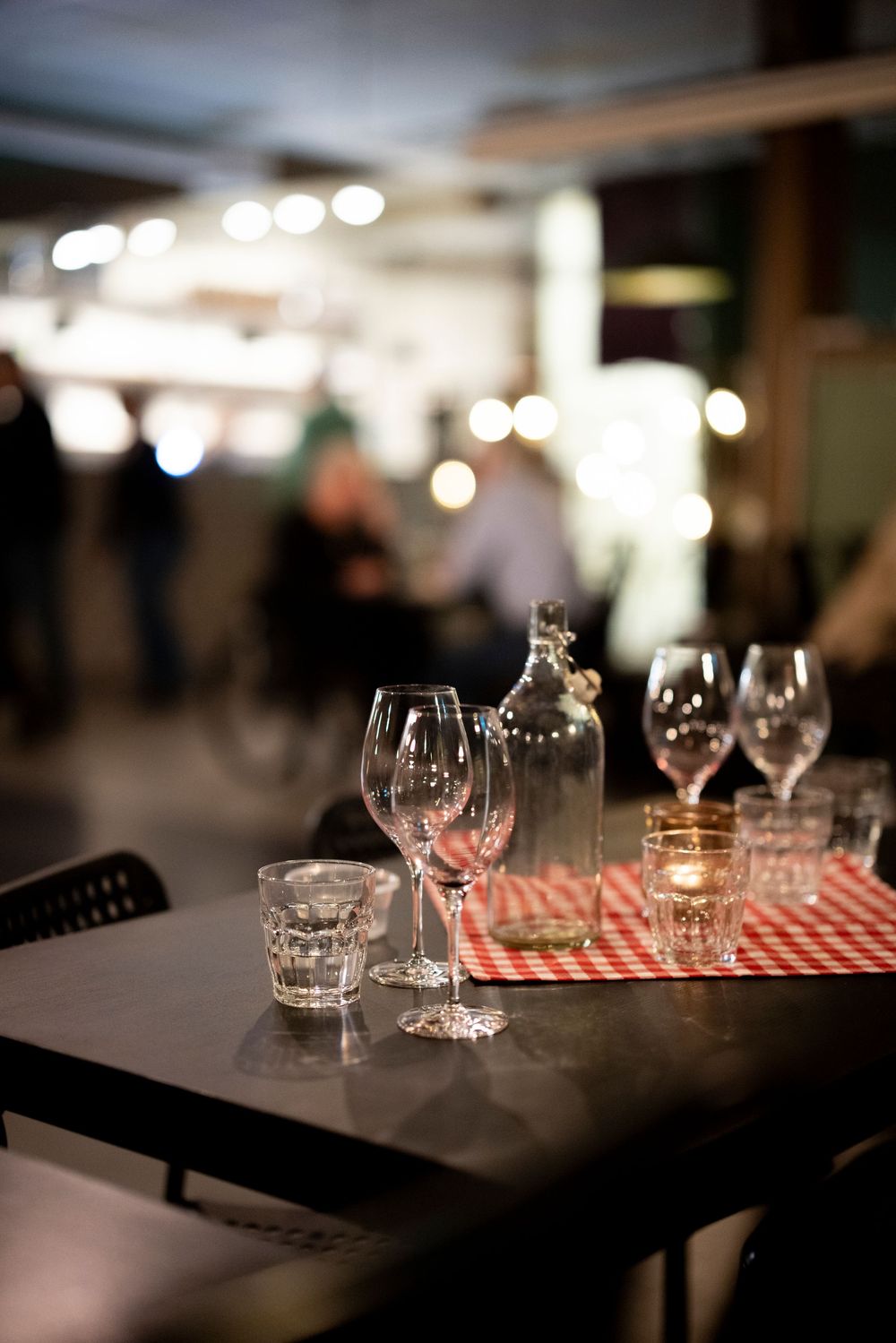 Restaurant table set with wine glasses, a carafe, and candles on a red checkered tablecloth in a busy dining room.