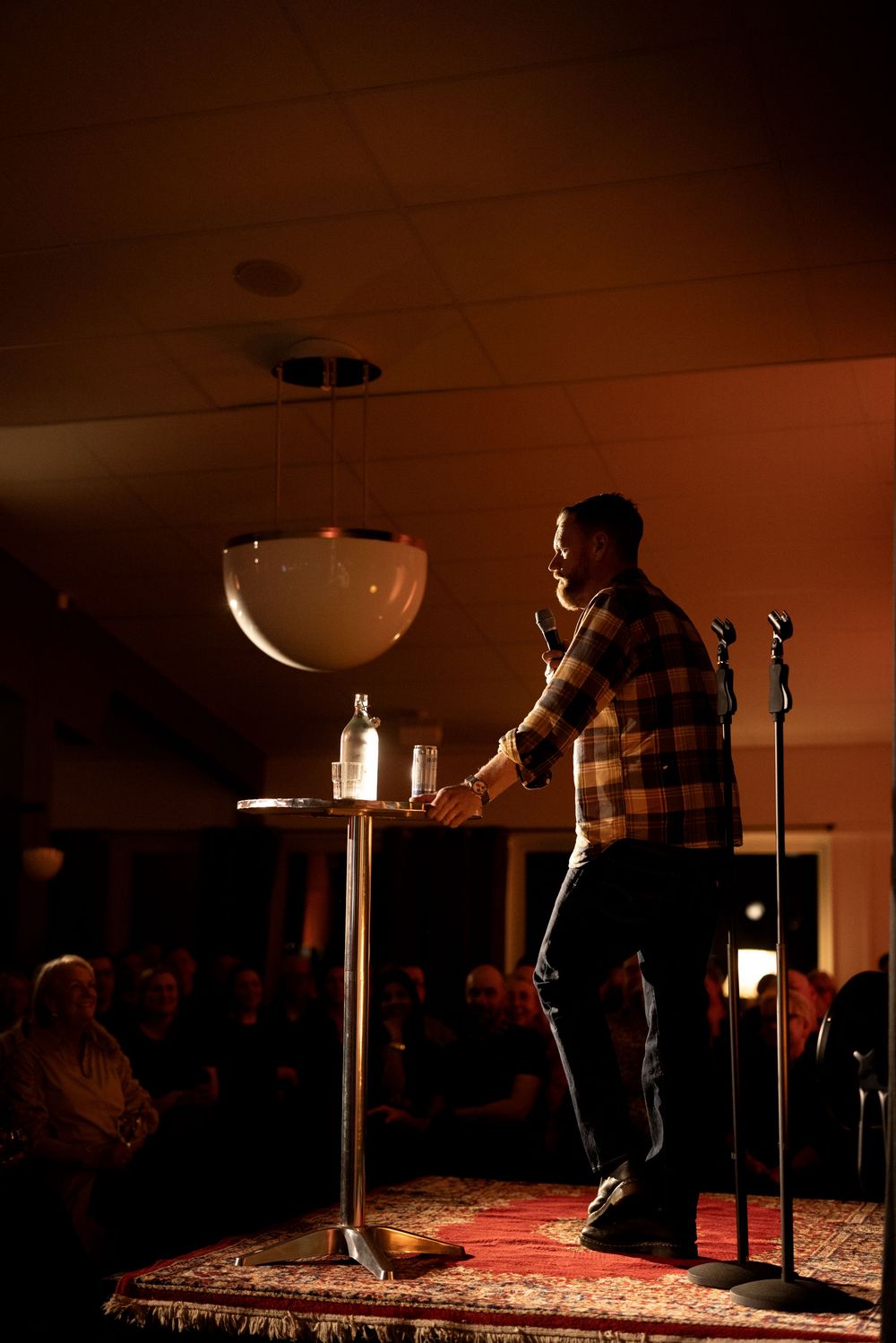Stand-up comedian performing on stage with microphone, warm lighting, and audience in background at comedy club.