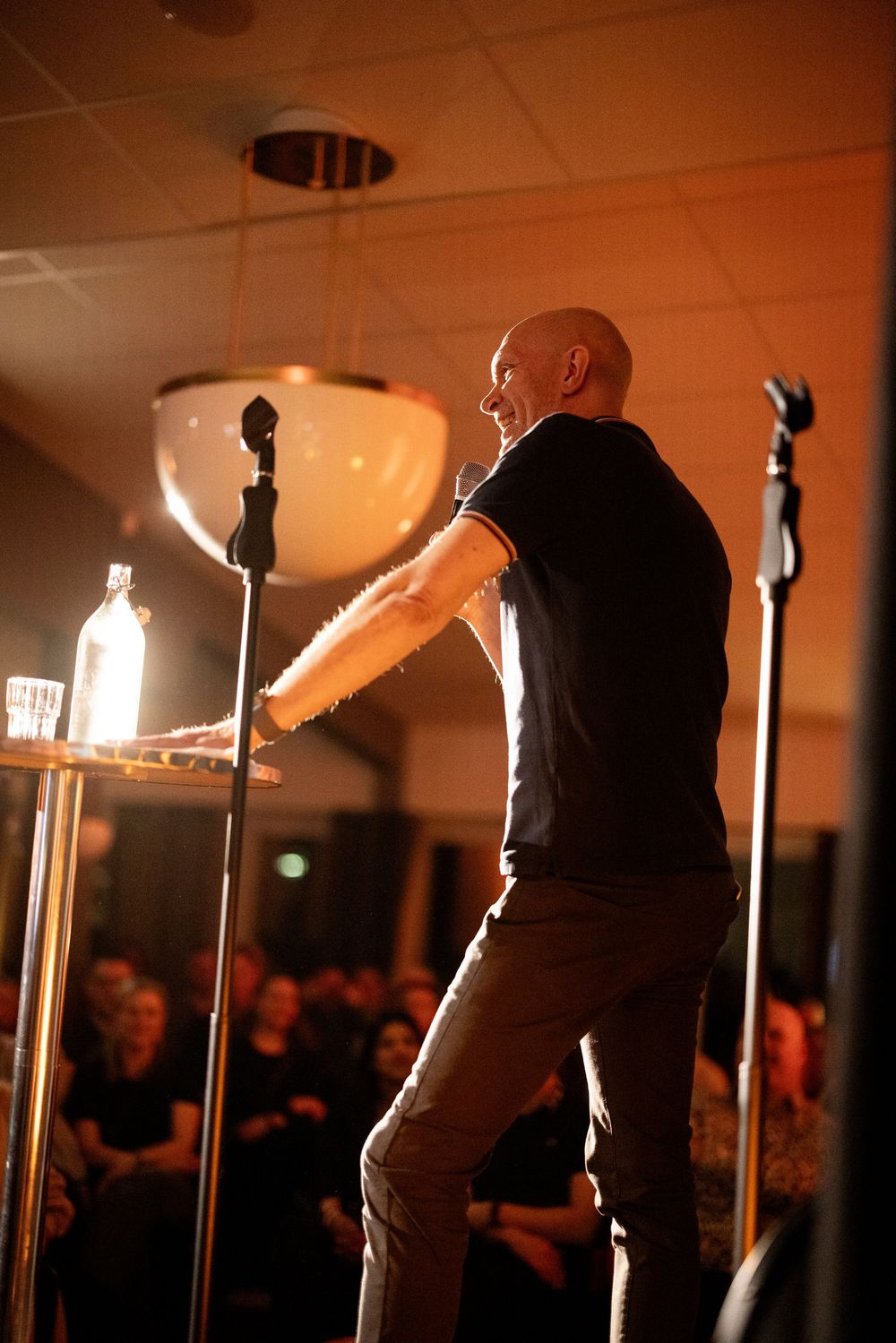 Bald man performing on stage with microphones, gesturing to an audience in a warmly lit venue.