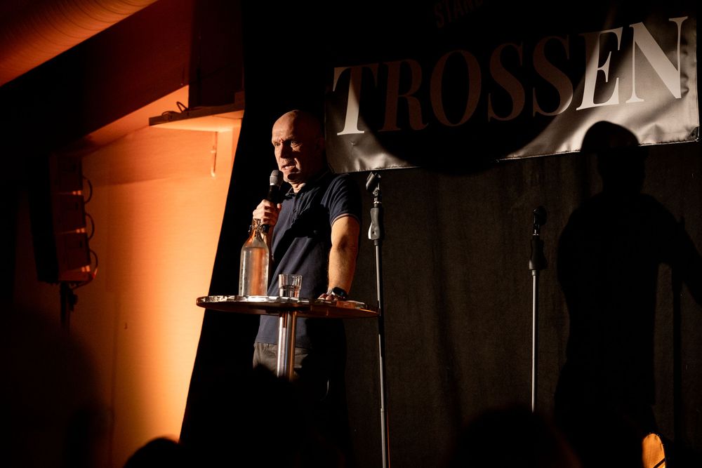 Man speaking on stage at Trossen venue, holding a bottle under dramatic spotlight lighting.