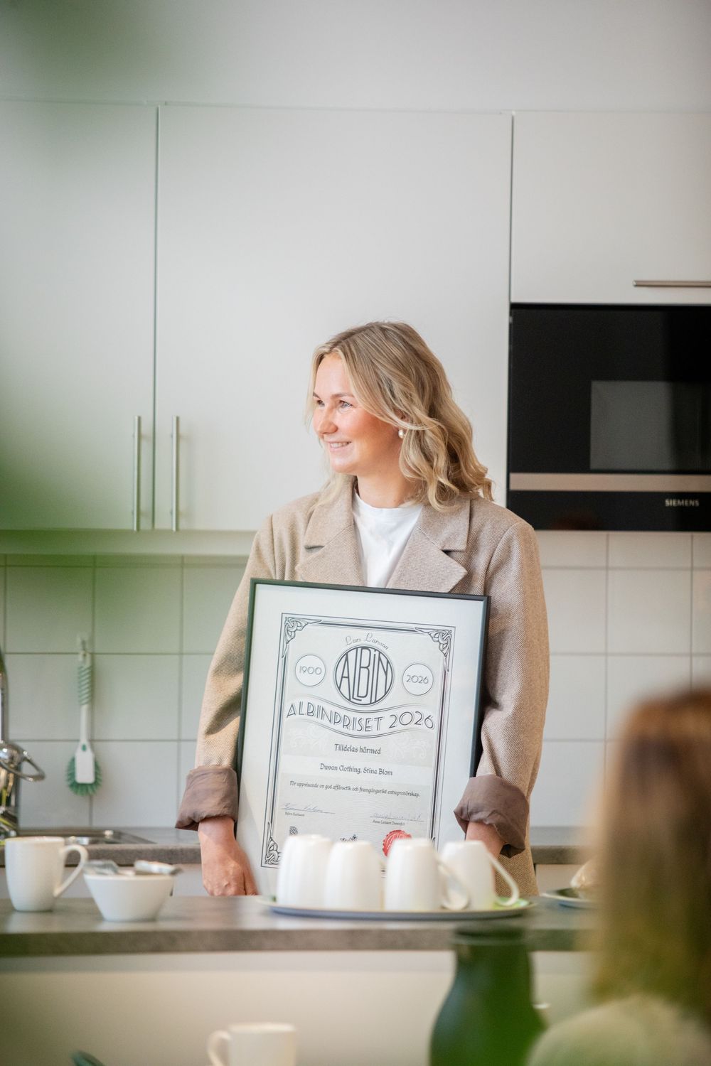 Smiling blonde woman holding a framed award certificate in a modern kitchen setting.