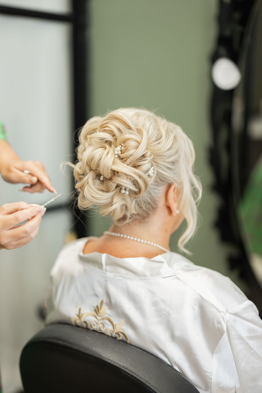 Hairstylist finishing elegant bridal updo on blonde woman wearing white robe and pearl necklace in salon.
