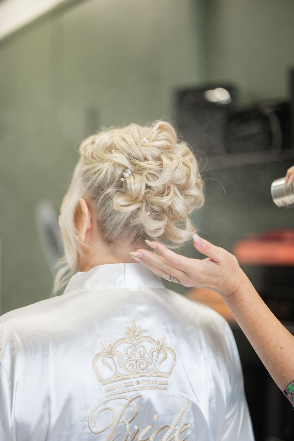 Bride in satin robe getting elegant braided updo hairstyle with hairspray at wedding preparation salon.