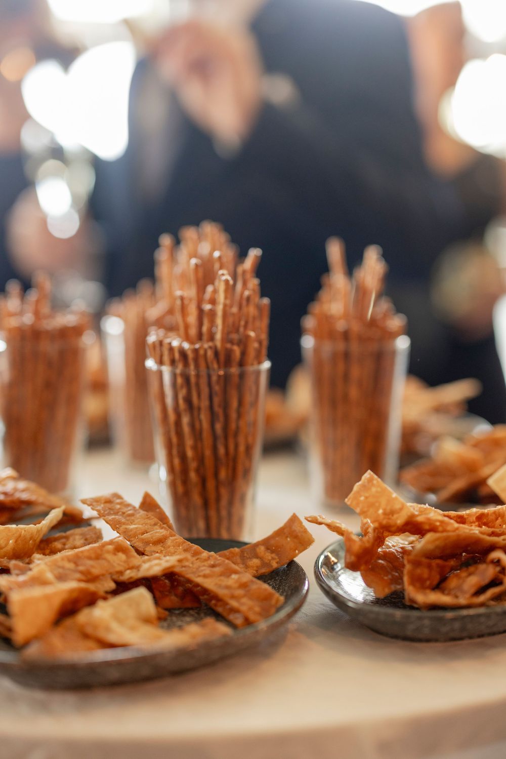 Assorted party snacks including pretzel sticks in glasses and crispy crackers on silver plates at a catered event.