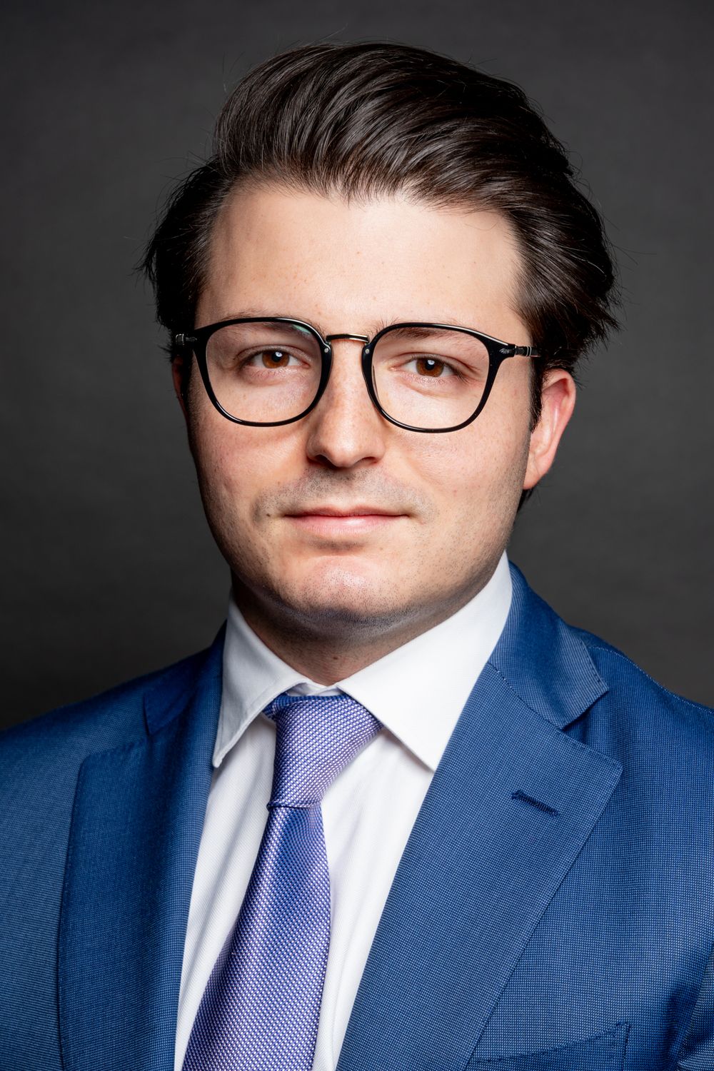 Professional man in a blue suit and glasses with a lavender tie on a gray background.