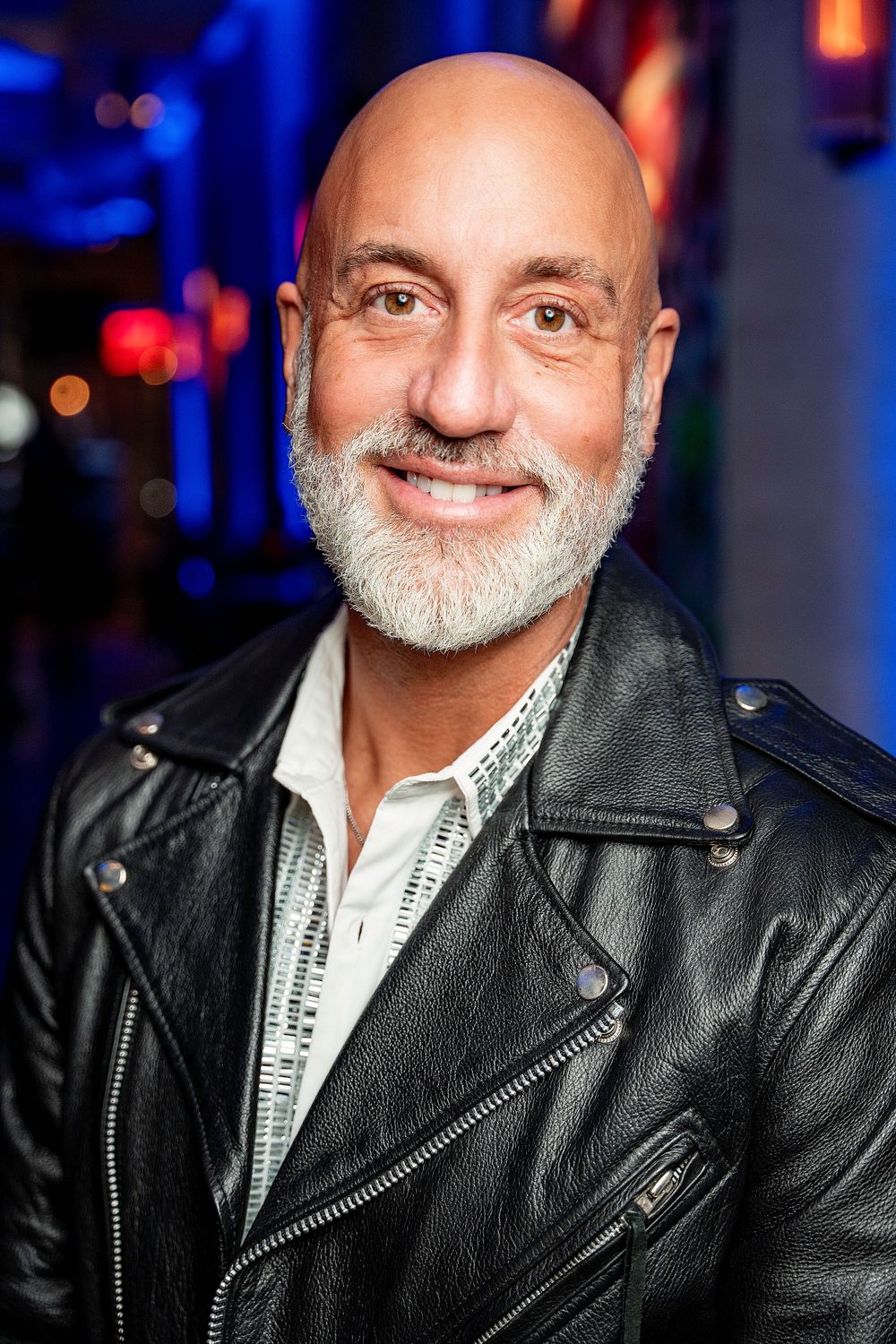 Bald man with a white beard wearing a black leather jacket smiling in a colorful venue.