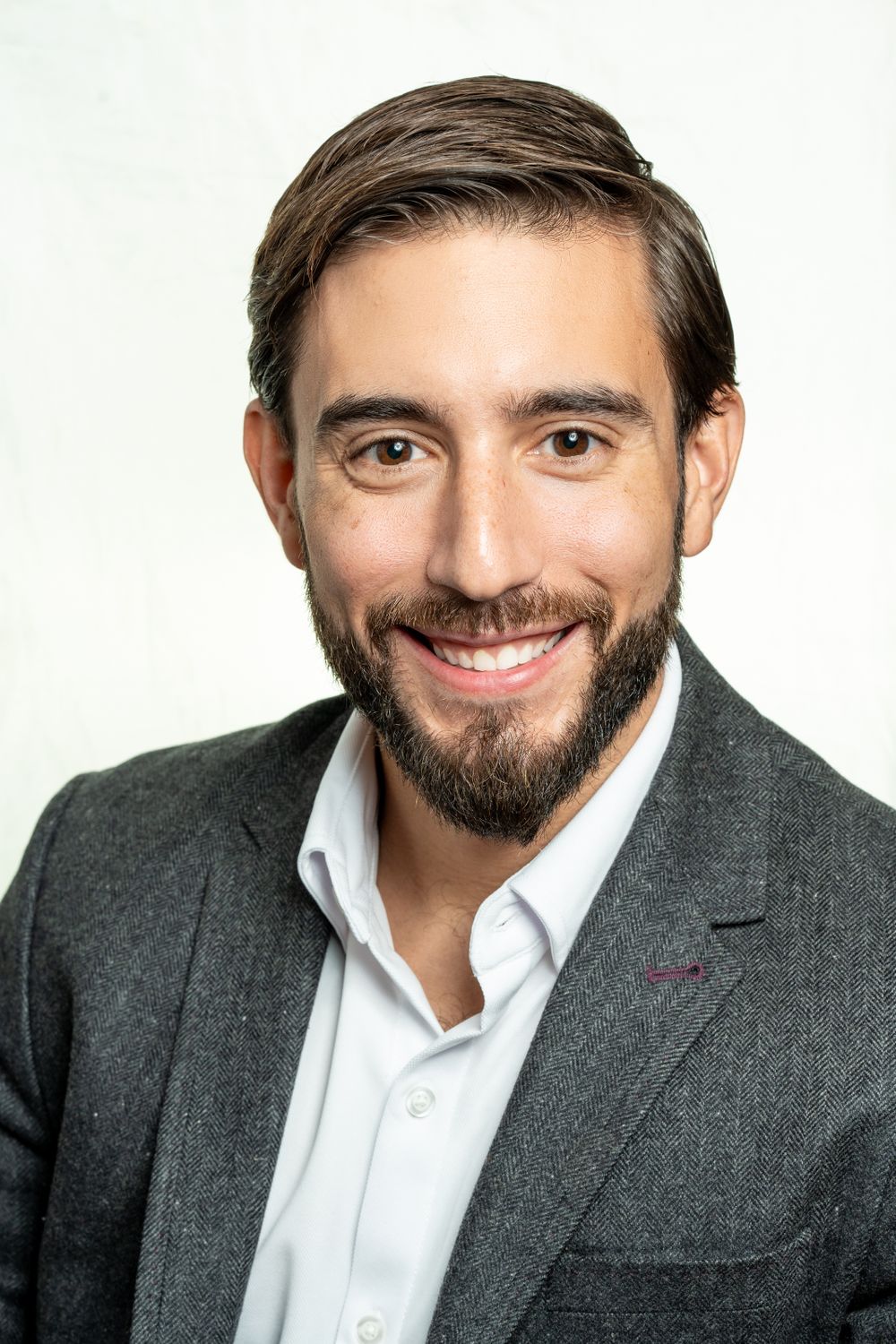 Smiling bearded man in a gray blazer and white shirt against a white background.