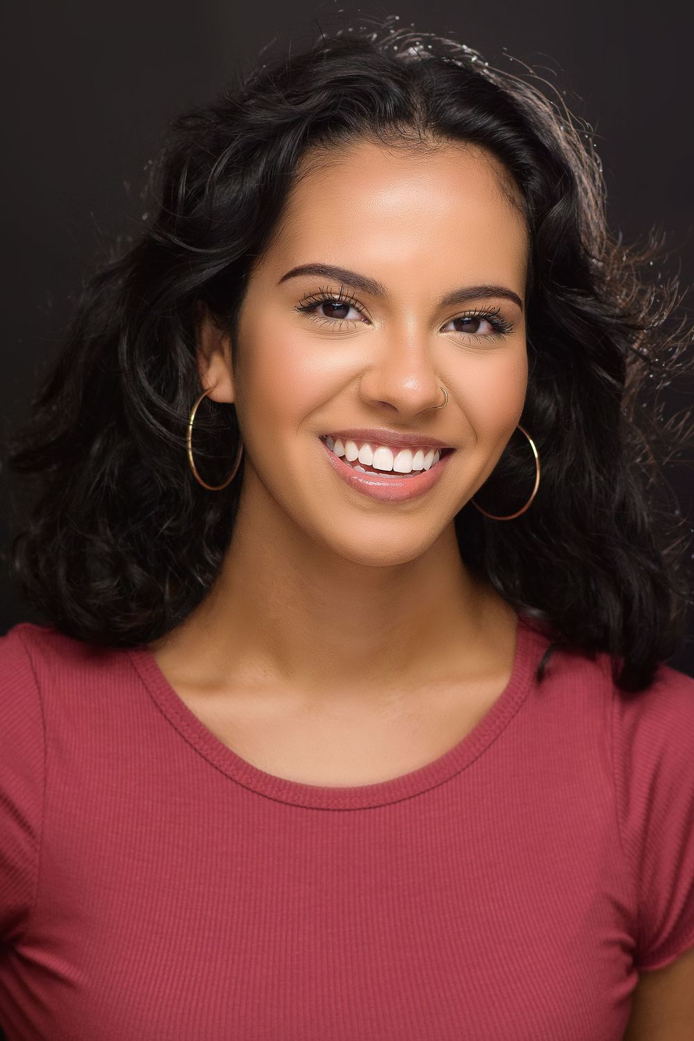 Young woman with curly dark hair smiling brightly, wearing gold hoop earrings and a pink top.