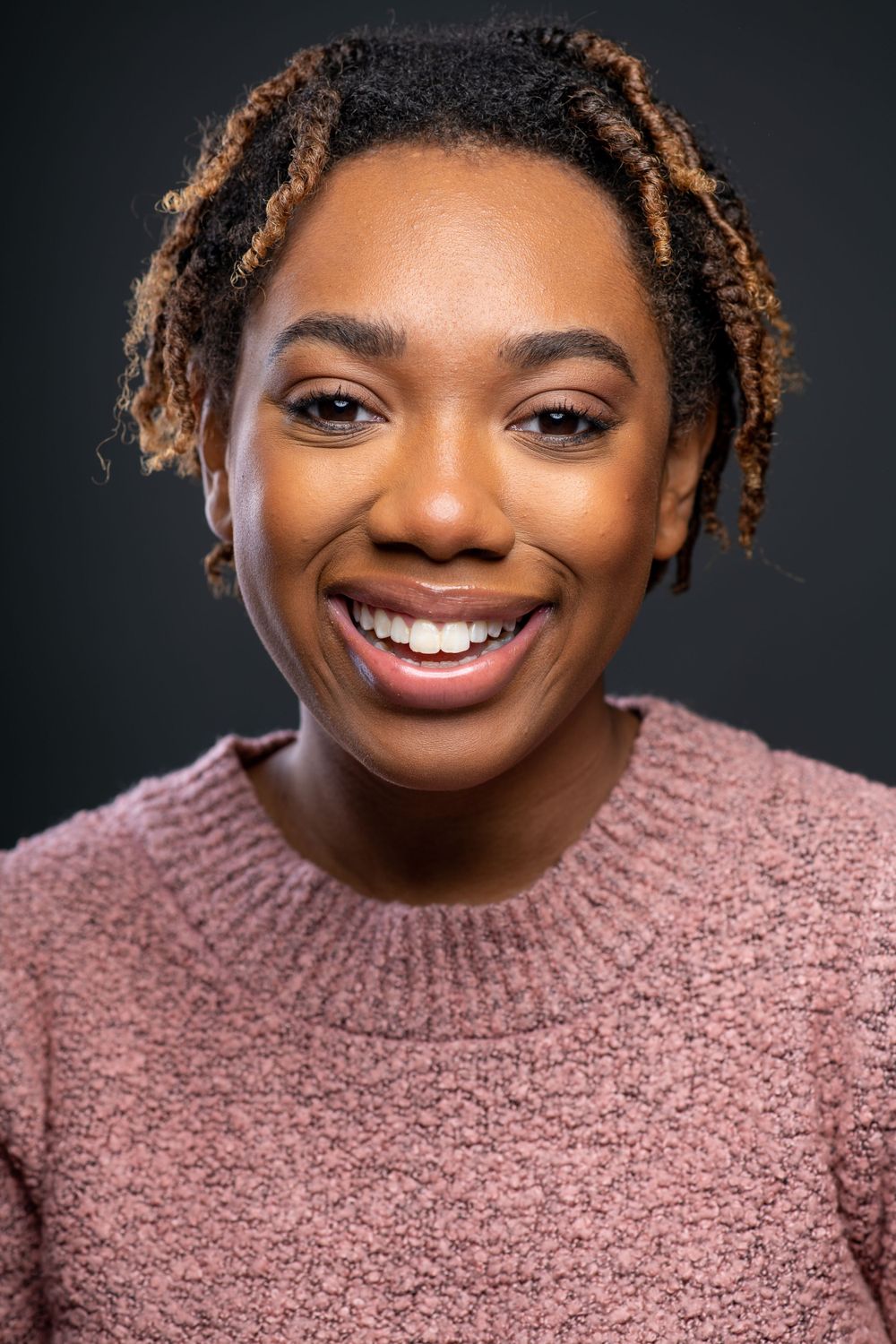 Joyful young woman with short dreadlocks smiling in a pink sweater on dark background.