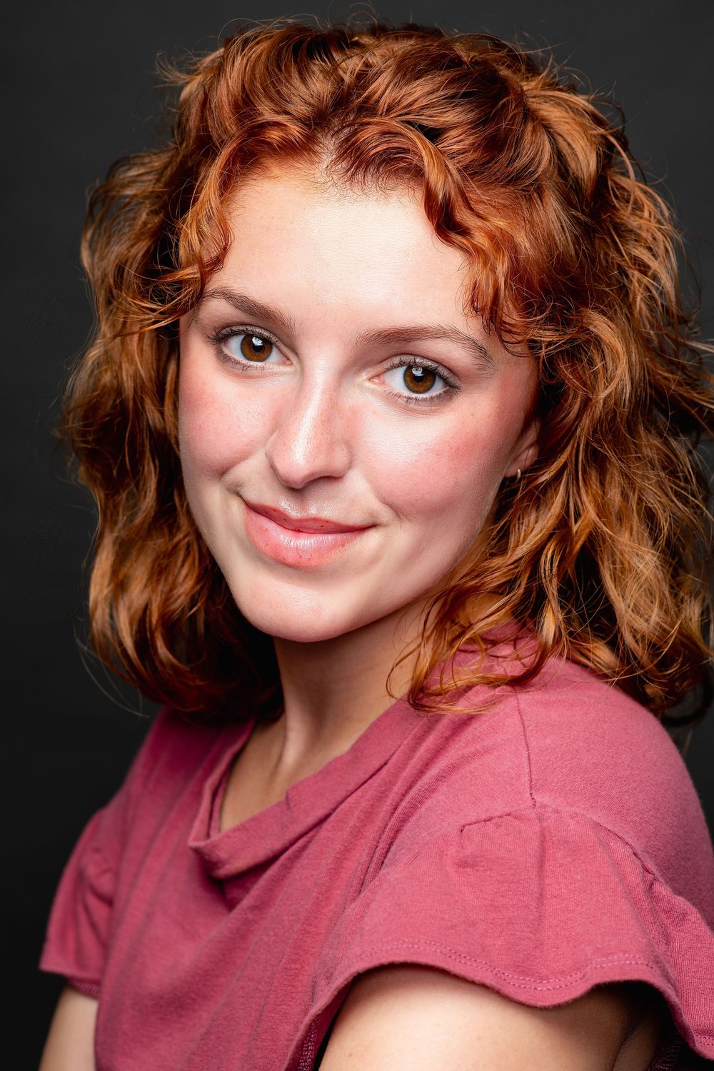 Young woman with curly red hair and freckles smiling softly in a pink top.