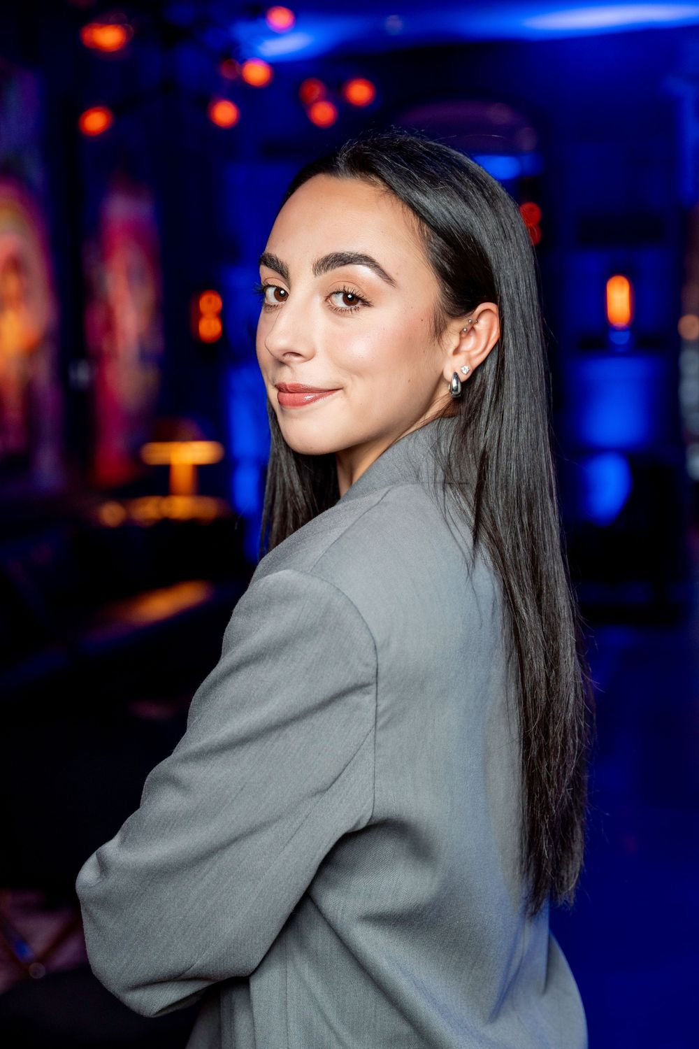 Young woman with straight dark hair in a gray blazer posing in a blue-lit venue.