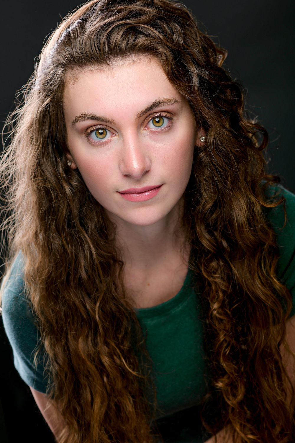 A young woman with long curly brown hair and green eyes poses for a professional headshot on dark background.