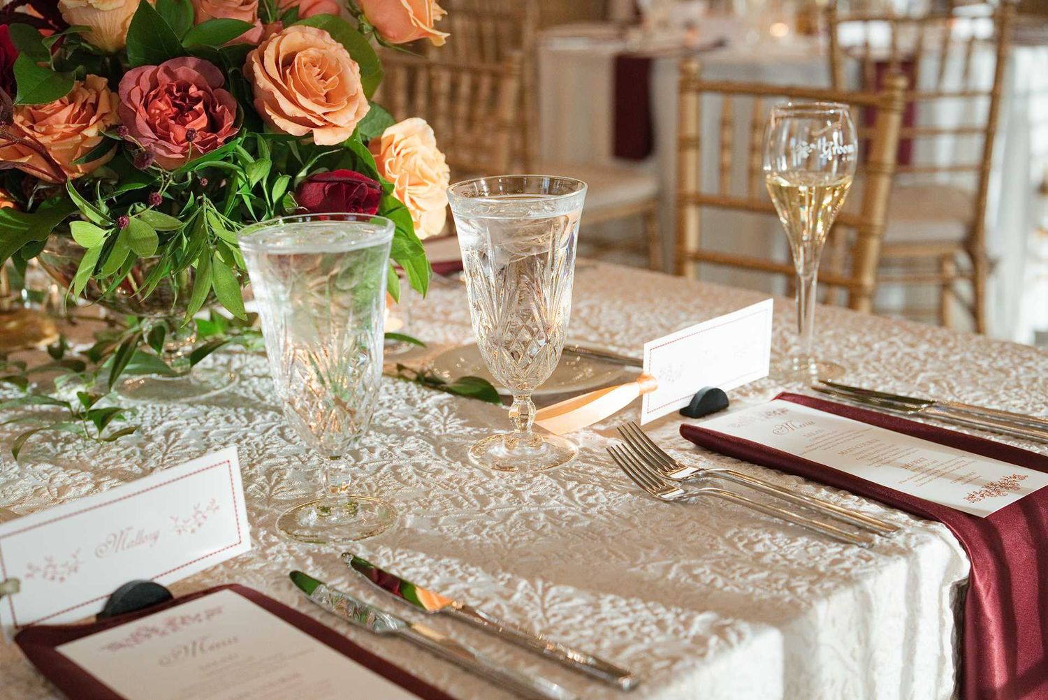 Elegant wedding reception table setting with crystal glassware, burgundy napkins, lace runner, and lush floral centerpiece.