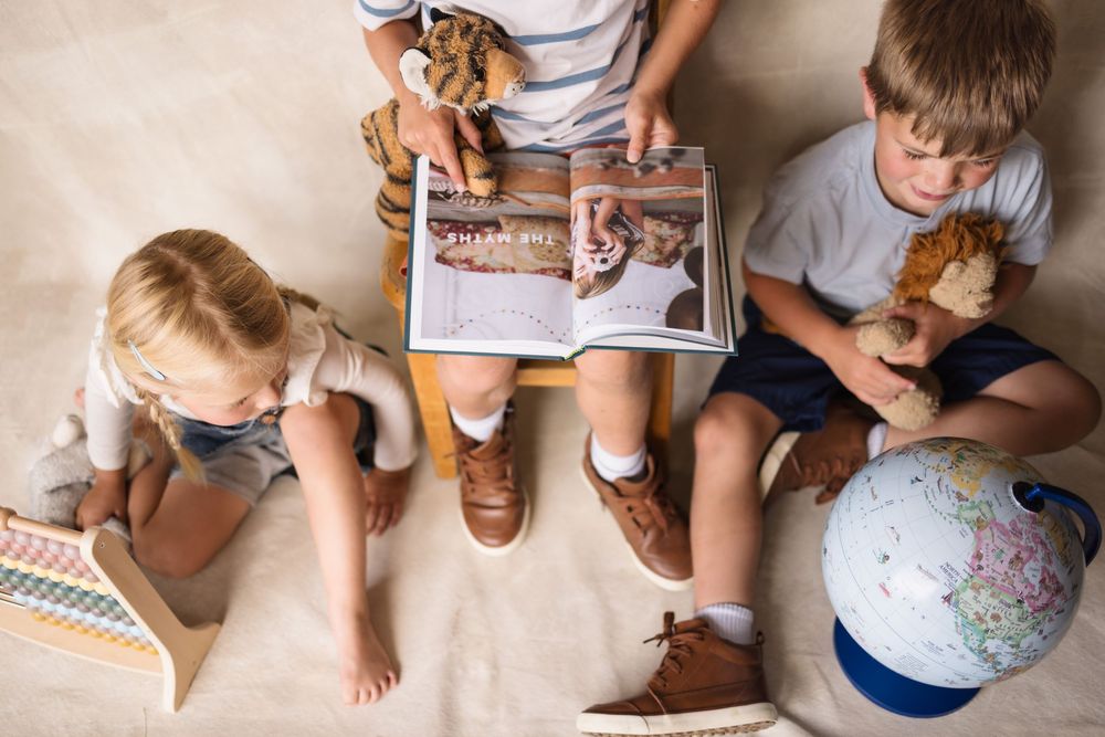 Three young children sit on floor reading a book with dinosaurs, holding stuffed animals and a globe nearby.