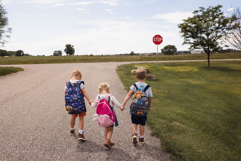 Three young children with backpacks walk hand-in-hand down a path toward a stop sign on a sunny day.