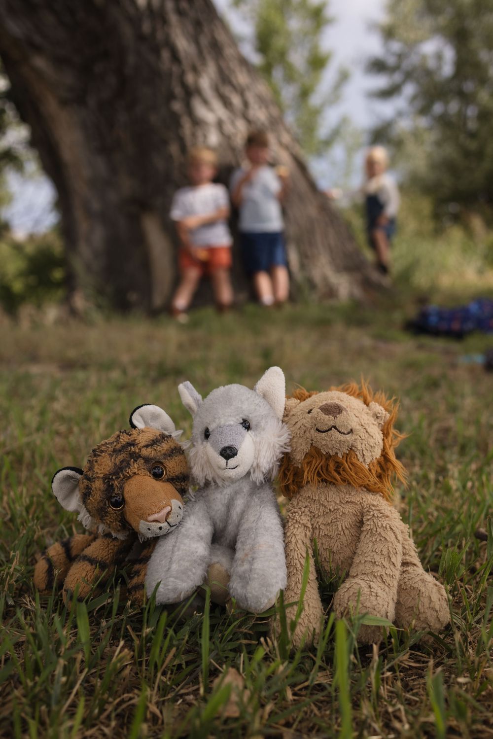 Three stuffed animal toys, tiger, wolf, and lion, sitting on grass with children playing near a tree in background.