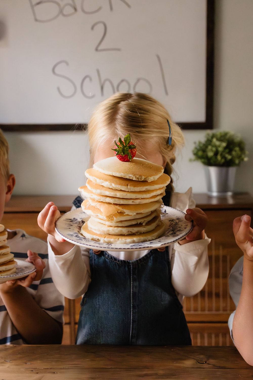 Girl holding tall stack of pancakes topped with strawberry in front of her face, Back 2 School sign in background.