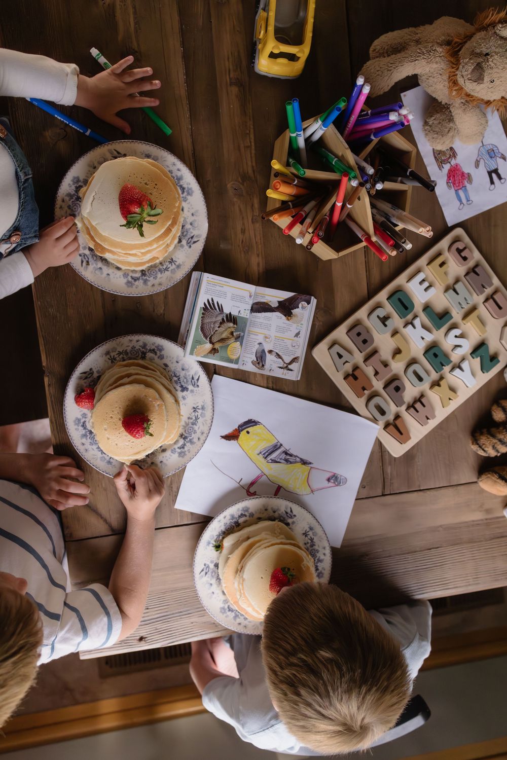 Overhead view of children eating strawberry pancakes at a wooden table with alphabet toys, colored pencils, and bird drawings.
