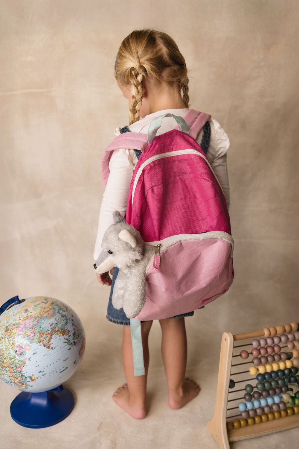 Young girl with blonde braids wearing a pink backpack with teddy bear, standing barefoot near a globe and abacus.