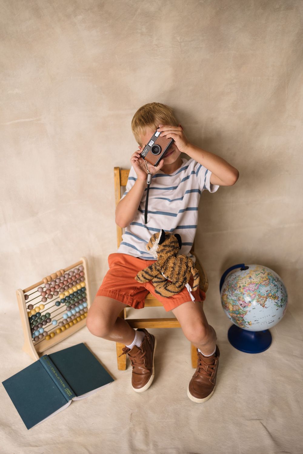 Young boy sitting on a chair holding a vintage camera, with a globe, abacus, and books around him.