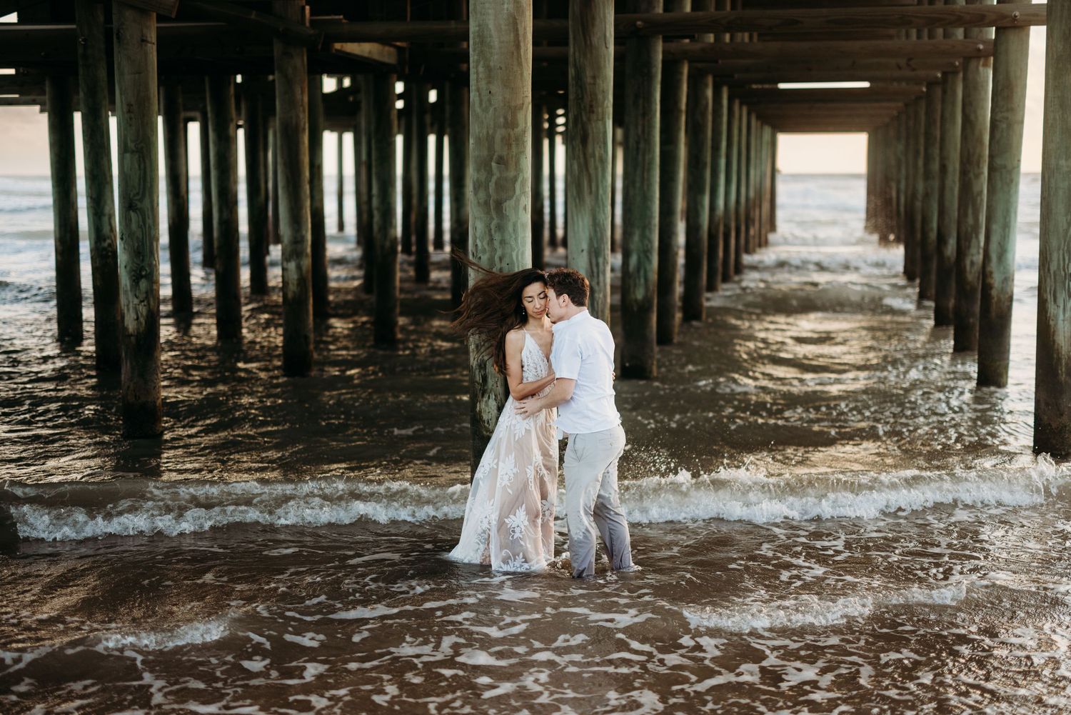 Couple embracing romantically under a pier with waves washing around their feet.