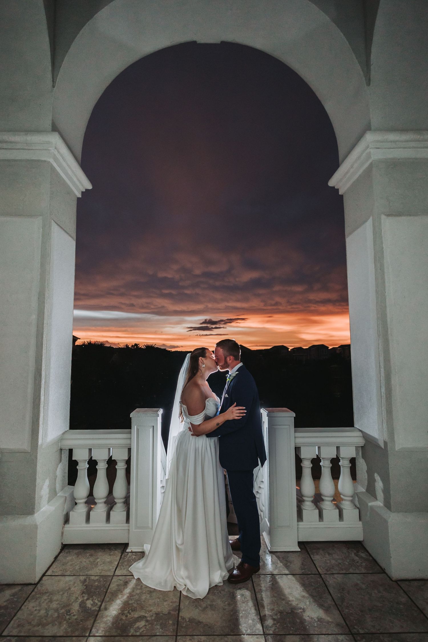 Bride and groom kissing under an arch at sunset, dramatic orange sky backdrop, elegant wedding venue balcony.
