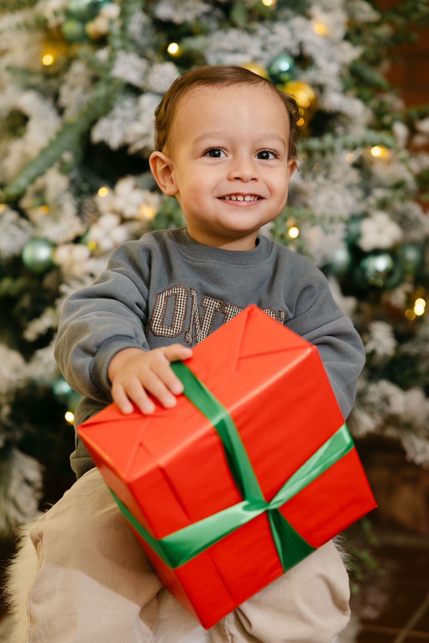 Smiling toddler boy holding a large red Christmas gift with green ribbon in front of a decorated holiday tree.