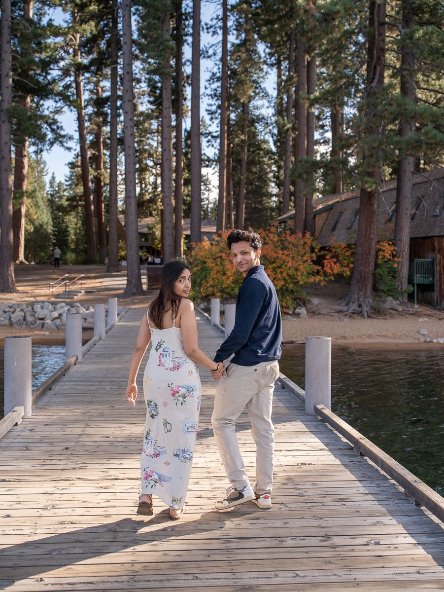 Couple embracing on a lakeside dock with a rustic boathouse and autumn foliage in the background.