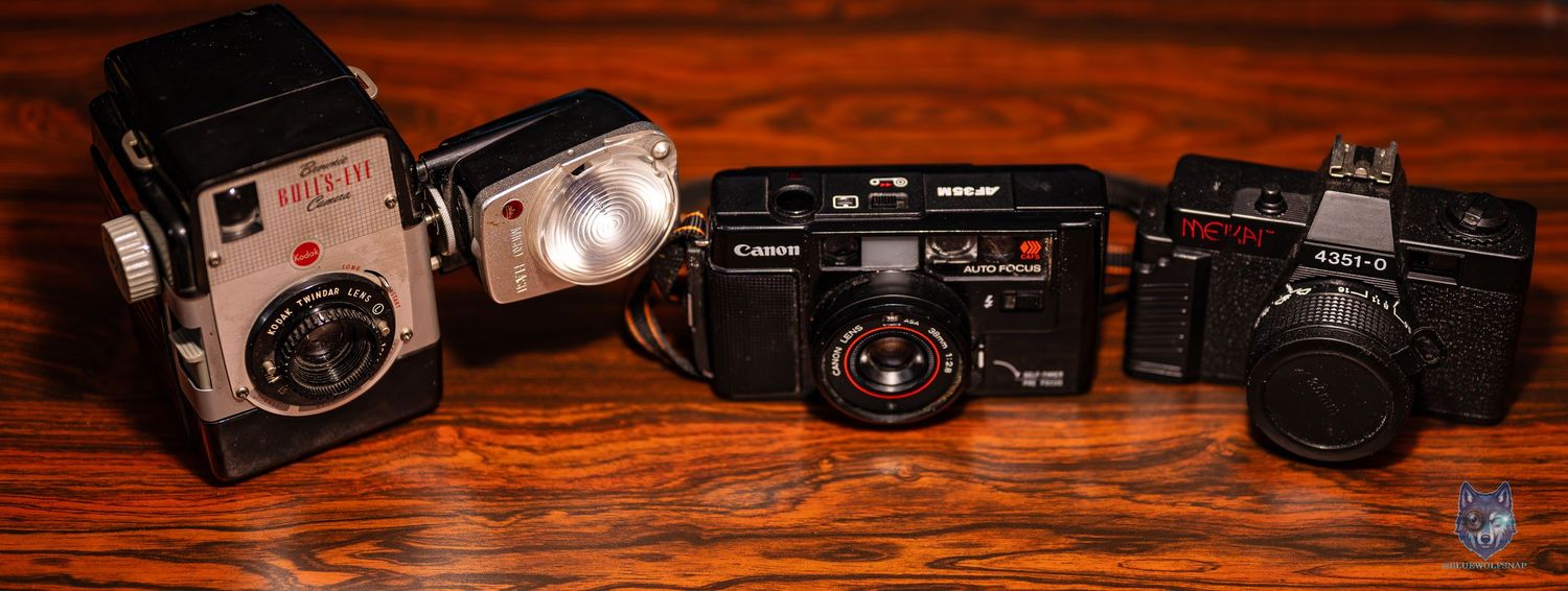 Four vintage film cameras displayed on a wooden surface, including a Holga, Canon, Rollei, and a compact point-and-shoot camera.