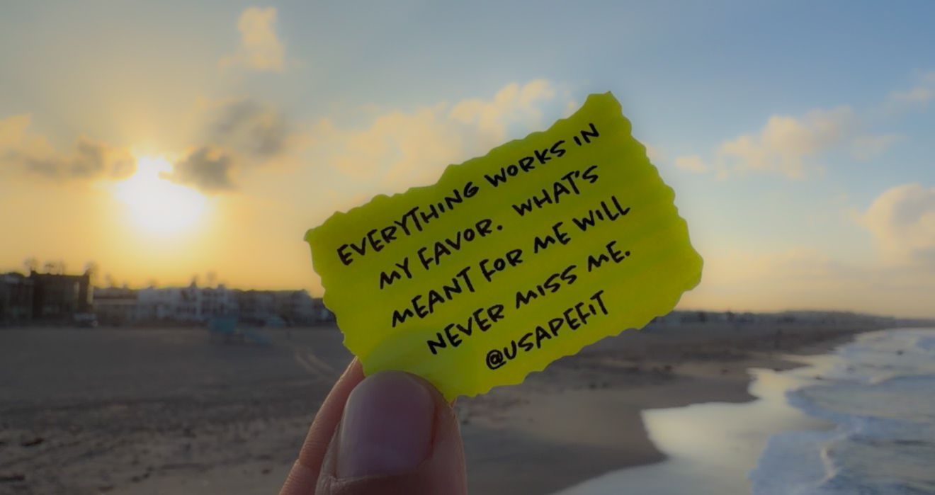 Hand holding yellow note with motivational quote on beach at sunset, reading Everything works in my favor @USAPER.