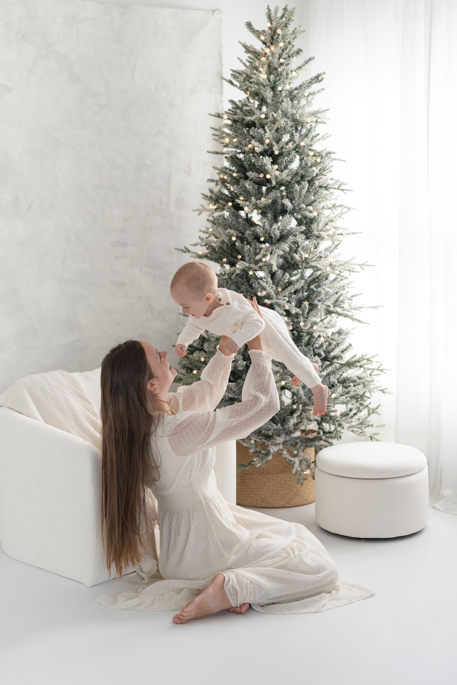 Portrait fine art de maman tenant bébé dans les airs assise au sol devant un sapin illuminé lors d'une séance photo de Noël au studio