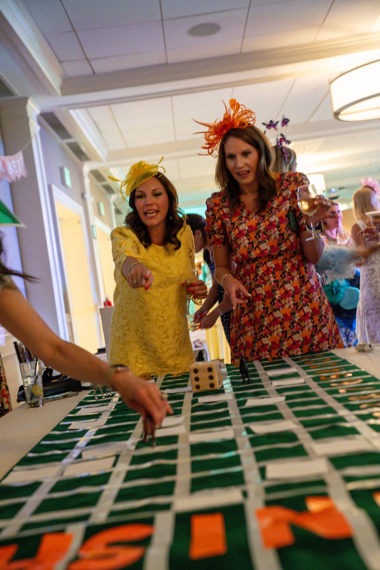 Two women in colorful dresses and fascinators playing a casino table game at an elegant Kentucky Derby party.