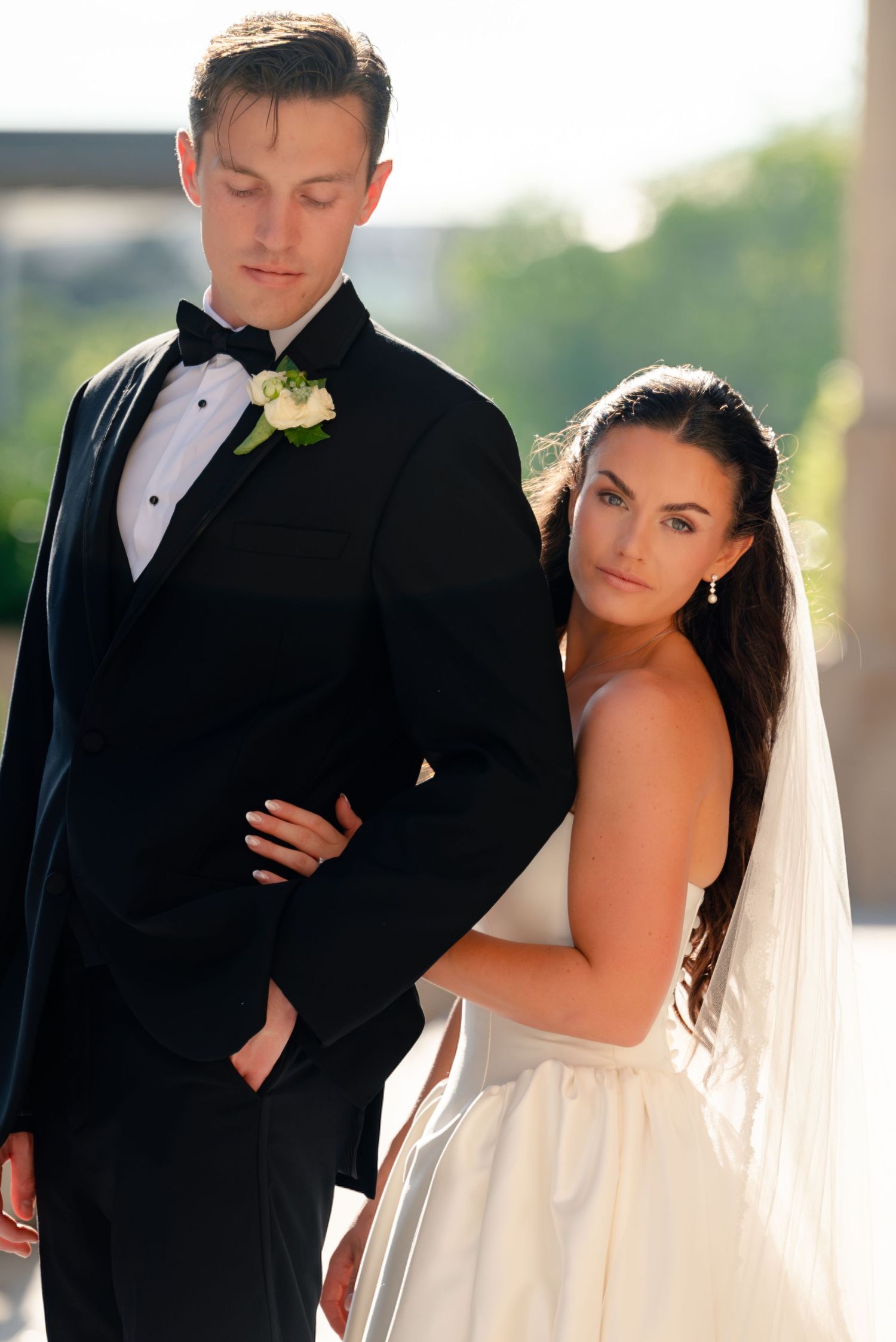 Bride in white gown and veil leans on groom in black tuxedo with boutonniere during outdoor wedding portrait.