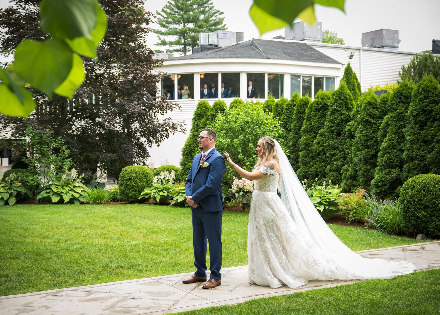 Bride in lace gown touches groom's shoulder during first look in lush garden outside elegant venue.