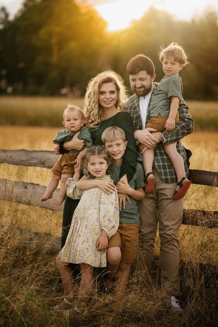 Family of six poses at golden hour by a rustic fence in a field, parents holding toddlers with children smiling warmly.