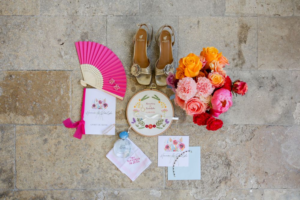 Colorful wedding flat lay with pink fan, gold heels, floral invitations, and vibrant orange, pink, and red roses on stone.