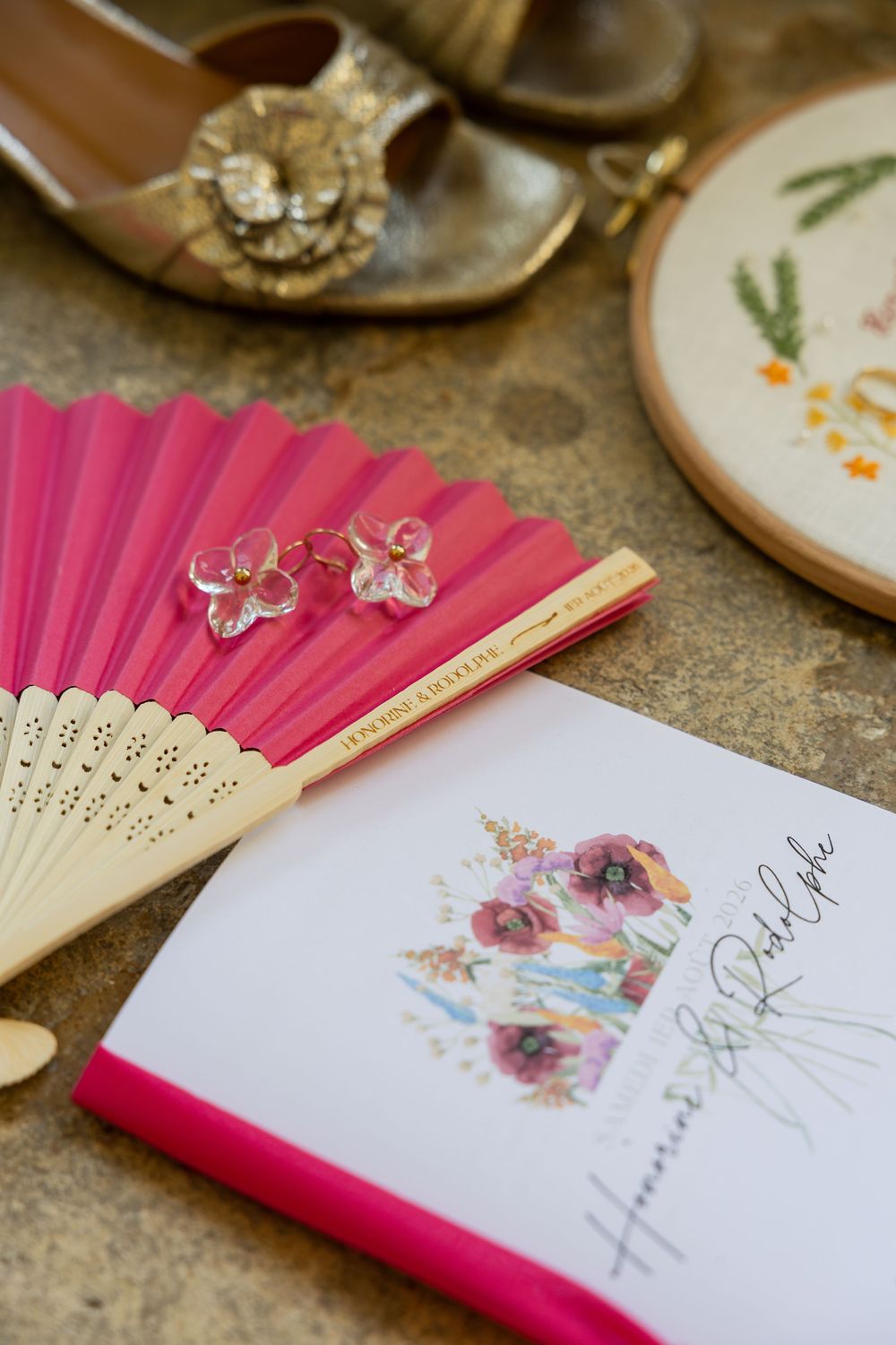 Pink folding fan, floral earrings, wedding program booklet, embroidery hoop, and gold bridal shoes arranged on a surface.
