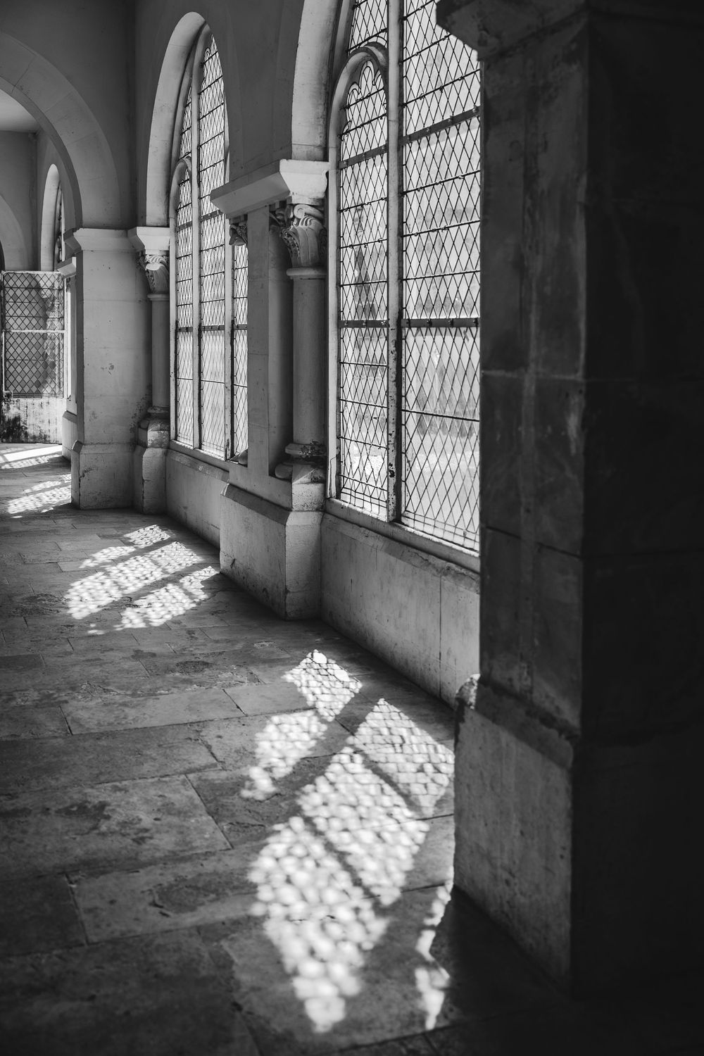 Sunlight filters through arched lattice windows casting geometric shadows on the stone floor of a historic corridor.