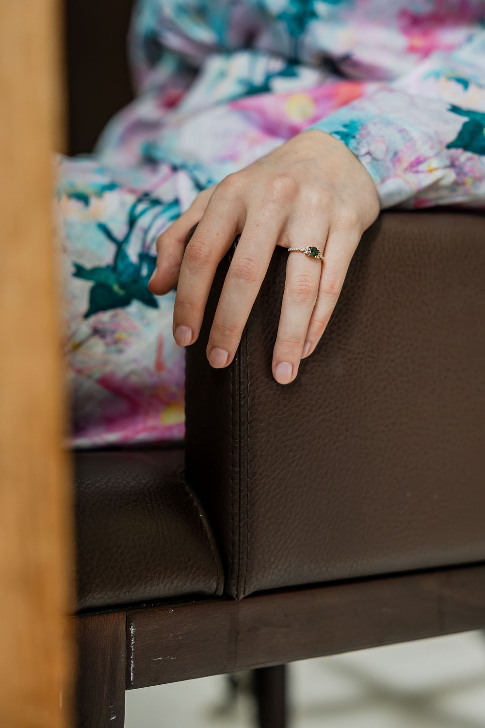 Close-up of a woman's hand with a gold ring resting on a dark brown leather chair, wearing a floral dress.