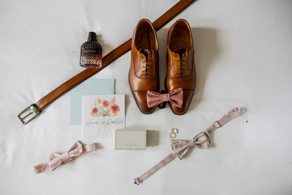 Brown oxford shoes, leather belt, pink bow ties, gold rings, cologne, and wedding stationery arranged on white fabric.