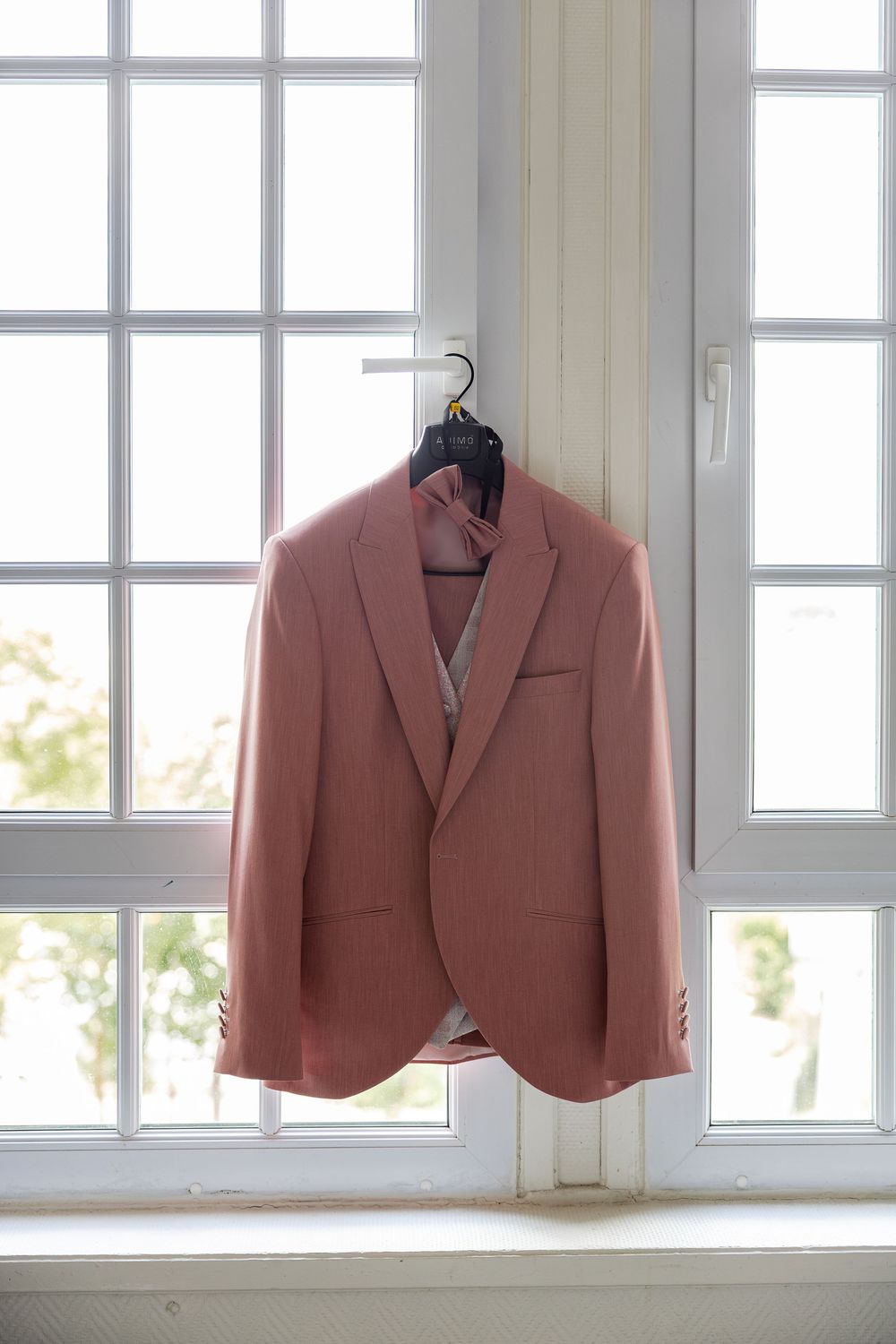 Pink men's suit jacket hanging on a hanger against a bright white window with natural light.