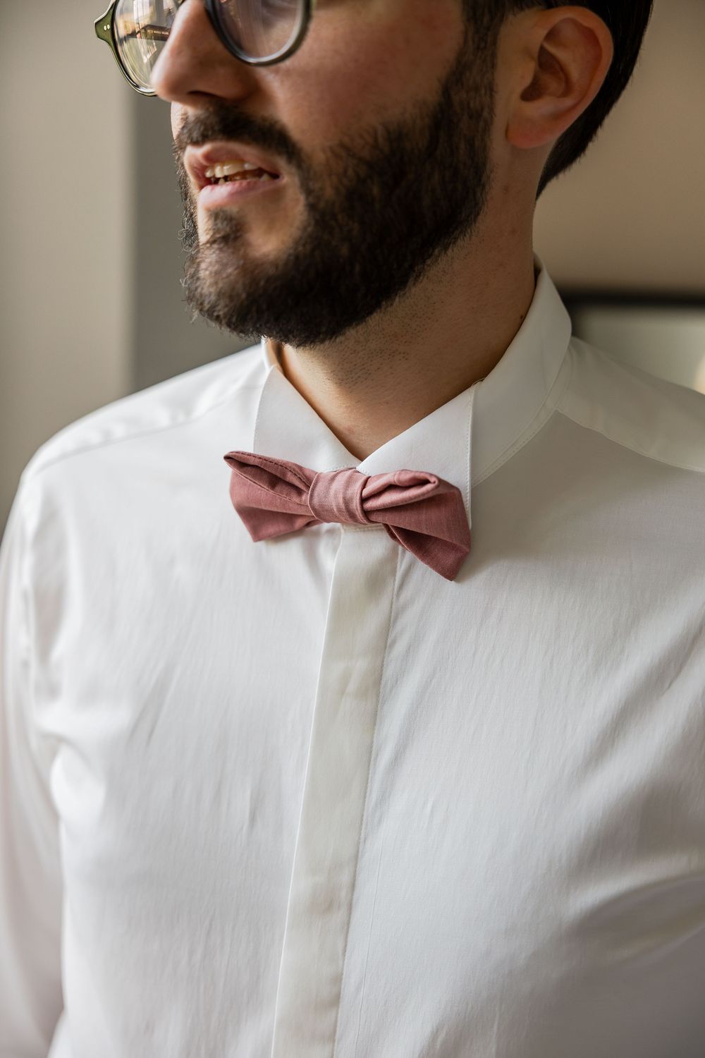 Smiling bearded groom in white dress shirt with pink dusty rose bow tie and glasses on wedding day.