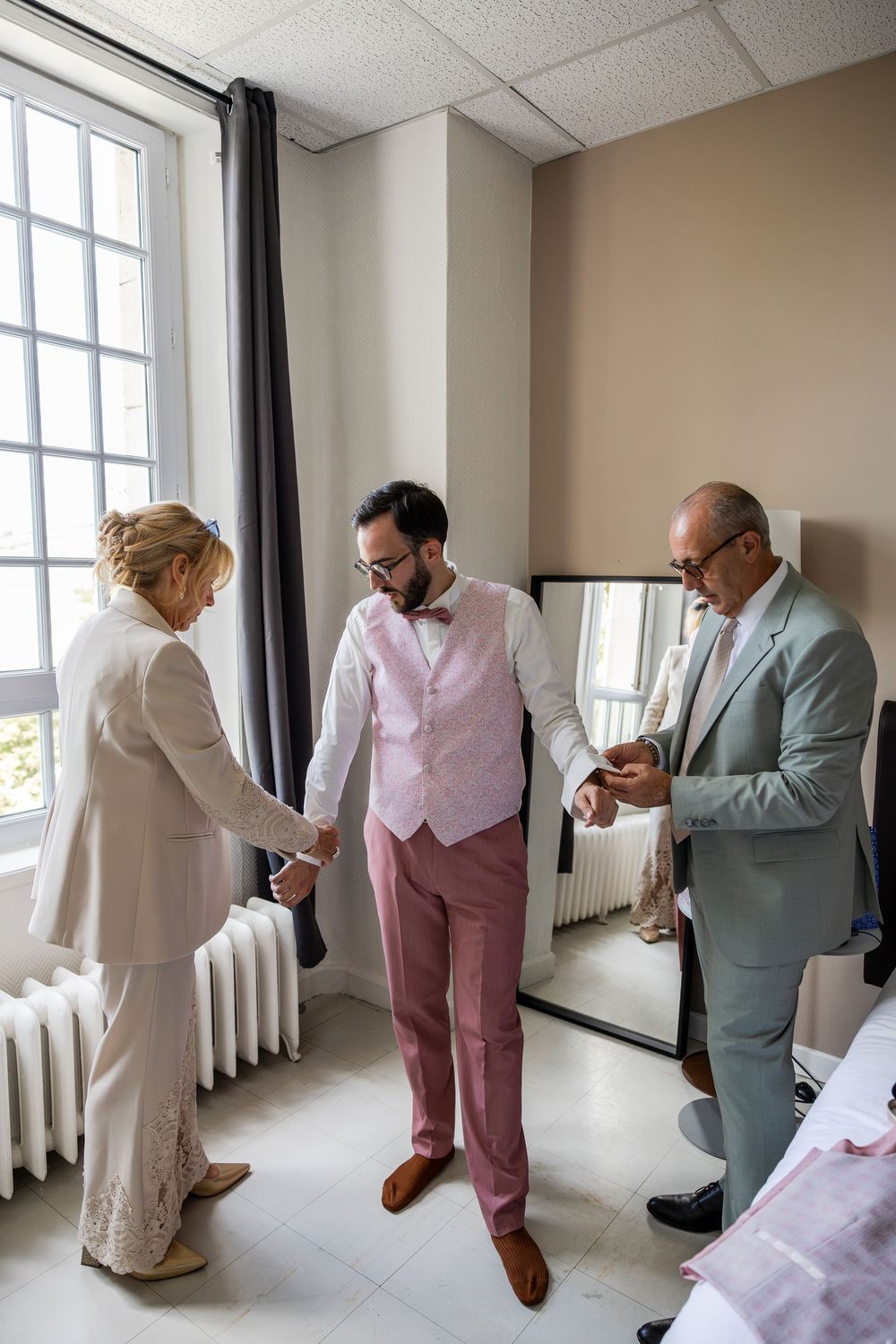 Groom in pink suit being helped by parents before wedding ceremony in bright room with large windows.
