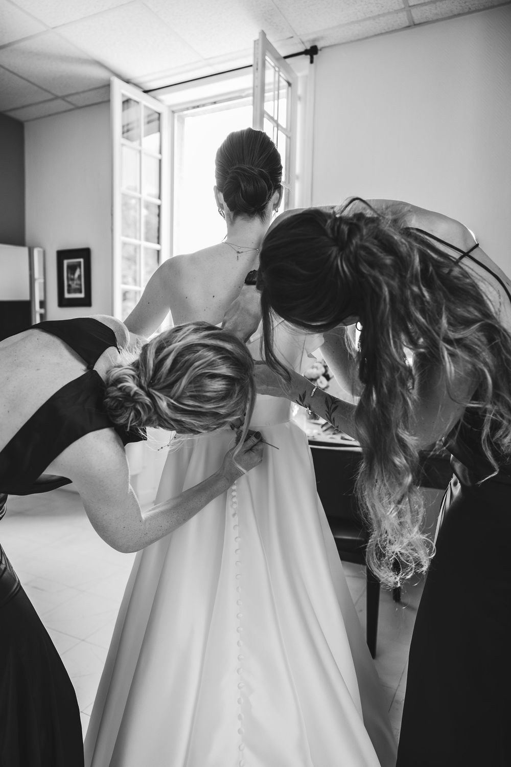 Black and white photo of bridesmaids helping bride button up wedding dress before ceremony.
