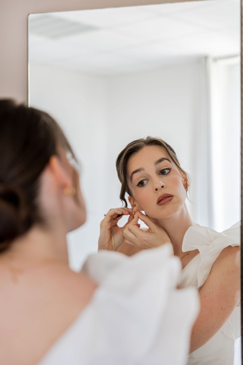 A bride in a white dress puts on earrings while gazing at her reflection in a large mirror.